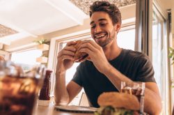 Happy young man eating a delicious burger at restaurant. Hungry young guy having stacked burger at fast food cafe.